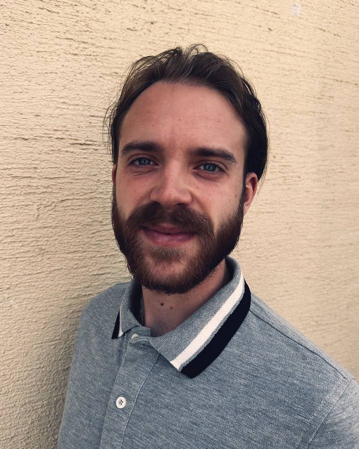 Portrait de Robin Sturmel : un homme aux cheveux bruns et à la barbe fournie, en polo gris à col rayé noir et blanc, devant un mur crépi beige.